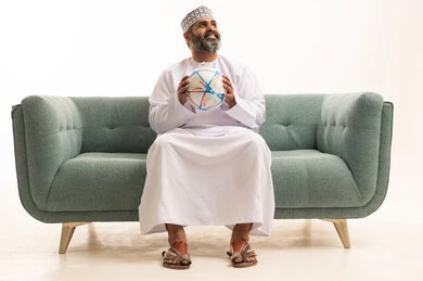 النظر الى شيء ما، أندية وتشجيع منتخبات, بورتريه لرجل عربي خليجي عماني يرتدي الدشداشة و الكمة يجلس على اريكه يقوم بالتفاعل والاندماج مع الفريق يمسك بيدية كره قدم , الشغف في متابعه الفرق، خلفية بيضاء