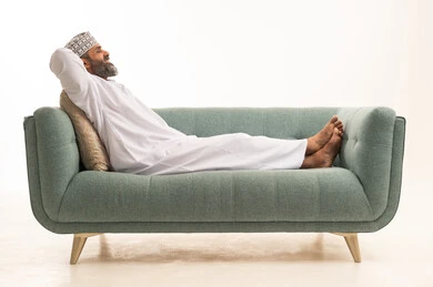 مفهوم الاسترخاء والاستمتاع بالأجواء الهادئة، بورترية لرجل عربي خليجي عماني يرتدي الدشداشة و الكمة يجلس على الاريكة ويضع يديه تحت رأسه، الشعور بالتعب والحاجة إلى النوم، ايماءات تدل على الراحة، خلفية بيضاء