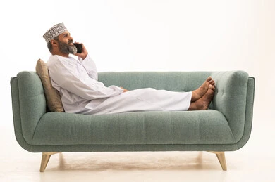 اجراء مكالمة هاتفية عبر الجوال، التواصل مع الاهل و الاصدقاء، بورترية لرجل عربي خليجي عماني يرتدي الدشداشة و الكمة يجلس على الاريكة يمسك بيدية الهاتف المحمول، خلفية بيضاء