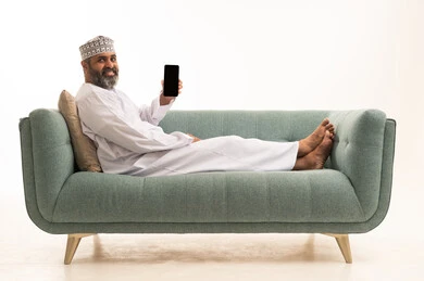 استخدام الاجهزة والتقنيات الحديثة المتطورة، النظر الى الكاميرا بتعابير السرور، عرض تطبيقات وبرامج الجوال، بورترية لرجل عربي خليجي عماني يرتدي الدشداشة و الكمة يجلس على الاريكة يقوم بعرض شاشة سوداء عبرالهاتف المحمول، خلفية بيضاء
