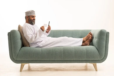 Payment and purchasing through mobile applications and programs, modern and advanced technologies, a portrait of an Arab Gulf Omani man wearing a dishdasha and kumma sitting on a sofa holding a mobile phone looking at a credit card, white background.