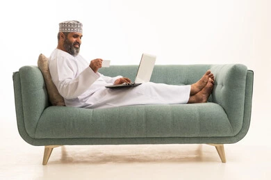 توفير خدمة الدفع الالكتروني لطلبات الاونلاين، بورترية لرجل عربي خليجي عماني يرتدي الدشداشة و الكمة يجلس على الاريكة يمسك بيده اللابتوب والبطاقة البنكية، خلفية بيضاء