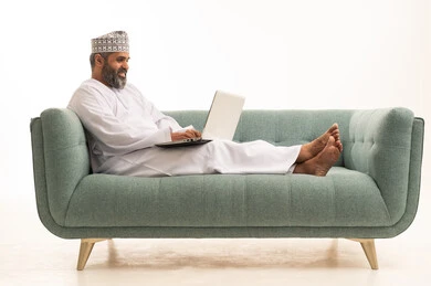 Using the computer to accomplish tasks, browsing the internet and social media programs, a portrait of an Arab Gulf Omani man wearing a dishdasha and kumma sitting on the sofa holding a laptop in his hand working on it, the development of technology and technical devices, white background.