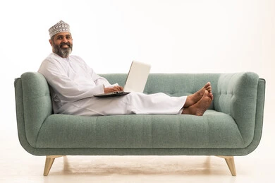 تصفح الانترنت وبرامج التواصل الاجتماعي, إستخدام الحاسوب لإنجاز المهام، بورترية لرجل عربي خليجي عماني يرتدي الدشداشة و الكمة يجلس على الاريكة يحمل بيده جهاز الاب توب المحمول يقوم بالعمل عليه، تطور التكنولوجيا والأجهزة التقنية، خلفية بيضاء