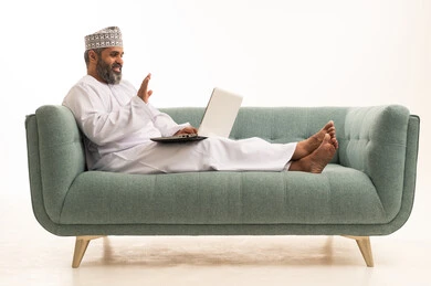 Expressions of peace and greeting, the development of technology and the use of modern technical devices, accomplishing work online through a laptop, a portrait of an Arab Gulf Omani man wearing a dishdasha and a kumma sitting on a sofa making a video call, white background.