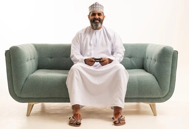 The concept of enjoyment and entertainment, gestures of interaction and engagement, a portrait of a Gulf Arab Omani man wearing a dishdasha and kumma sitting on the sofa with expressions of joy, having a good time playing video games, enjoying modern technologies and gadgets, white background.