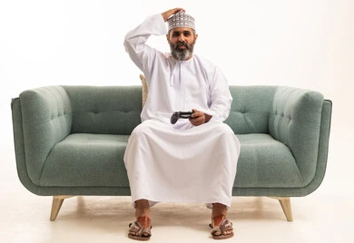 اللعب بألعاب الفيديو الترفيهية، تعابير الحزن والخسارة, استخدام الأجهزة التكنولوجية والتقنيات الحديثة،  بورترية لرجل عربي خليجي عماني يرتدي الدشداشة و الكمة يجلس على الاريكة يمسك بيده عصا التحكم يضع يده على رأسه بإيماءات الحسرة، خلفية بيضاء