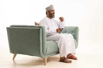 Expressions of victory and triumph, interaction and immersion with PlayStation games, modern electronic entertainment technologies, and a portrait of an Arab Gulf Omani man wearing a dishdasha and kumma sitting on the couch holding a game controller playing video games, white background.