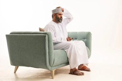 Expressions of sadness and loss, the use of technological devices and modern techniques, a portrait of an Arab Gulf Omani man wearing a dishdasha and kumma sitting on the sofa holding a game controller with his hand on his head in gestures of regret, playing recreational video games, white background.