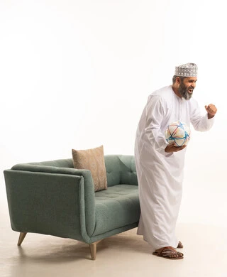 الشغف بمباريات كرة القدم، الانسجام والتفاعل مع المنافسة, رفع اليد الى الاعلى بتعابير الفوز والانتصار, بورترية لرجل عربي خليجي عماني يرتدي الدشداشة و الكمة يقف بجانب الاريكة يمسك الكرة, خلفية بيضاء