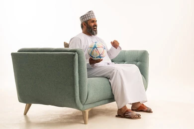 رفع اليد الى الاعلى بتعابير الفوز والانتصار، الانسجام والتفاعل مع المنافسة, الشغف بمباريات كرة القدم, بورترية لرجل عربي خليجي عماني يرتدي الدشداشة و الكمة يجلس على الاريكة يمسك الكرة, خلفية بيضاء