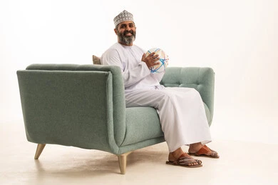 Integration through encouraging football, the passion for football matches, a portrait of an Arab Gulf Omani man wearing a dishdasha and kumma sitting on a sofa holding a ball, white background.
