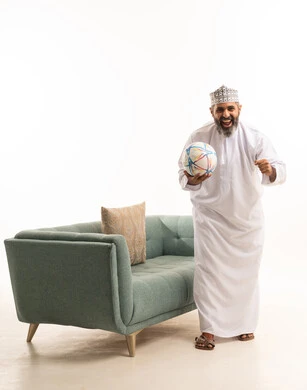 Harmony and interaction with competition, passion for football matches, raising the hand up with expressions of victory and triumph, a portrait of an Arab Gulf Omani man wearing a dishdasha and kumma standing next to the sofa holding a ball, white background.