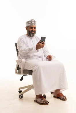 Browsing social media, a portrait of an Arab Gulf Omani man wearing a dishdasha and kumma sitting on a chair using a mobile phone, talking and chatting with friends via mobile, the concept of comfort and sitting, white background.