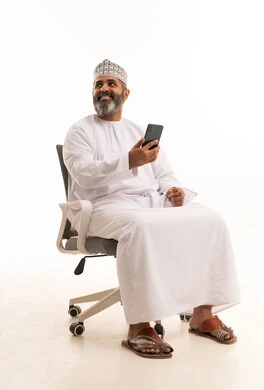 Looking at something, browsing social media, a portrait of an Arab Gulf Omani man wearing a dishdasha and kumma sitting on a chair using a mobile phone, talking and chatting with friends via mobile, the concept of comfort and sitting, white background.