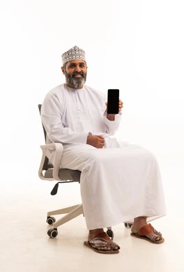 A display of a blank black screen, the use of modern technologies, the quality of mobile applications and services, a portrait of an Arab Gulf Omani man wearing a dishdasha and kumma sitting on a chair looking at the camera with gestures of joy, white background.