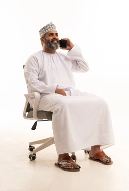 Making a phone call via mobile, chatting with family and friends, a portrait of an Arab Gulf Omani man wearing a dishdasha and a kumma sitting on a chair and holding a mobile phone in his hand, white background.