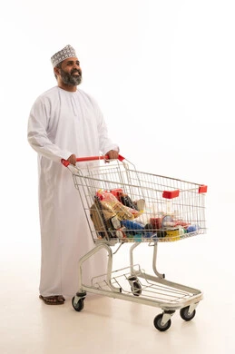 اقتناء المستلزمات اللازمة، الاستفادة من الخصومات على المنتجات، الاستمتاع بالتسوق في المولات والمراكز التجارية، بورتريه لرجل عربي خليجي عماني يرتدي الدشداشة و الكمة يجر عربة التسوق أمامه، الجسم بالطول الكامل، خلفية بيضاء