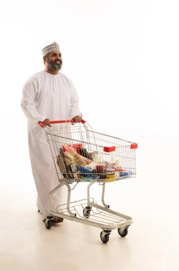 شراء الحاجيات شراء الحاجيات الضرورية، الاستفادة من الخصومات على المنتجات، الاستمتاع بالتسوق في المولات والمراكز التجارية، بورتريه لرجل عربي خليجي عماني يرتدي الدشداشة و الكمة يجر عربة التسوق أمامه، الجسم بالطول الكامل، خلفية بيضاء