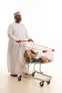 اقتناء الحاجيات الأساسية، الاستفادة من الخصومات على المنتجات، الاستمتاع بالتسوق في المولات والمراكز التجارية، بورتريه لرجل عربي خليجي عماني يرتدي الدشداشة و الكمة يجر عربة التسوق أمامه، الجسم بالطول الكامل، خلفية بيضاء