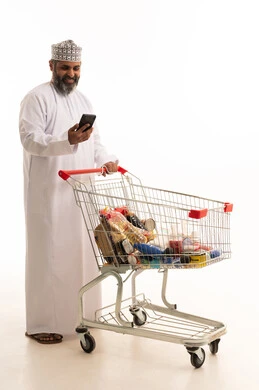 Browsing social media, taking advantage of discounts and sales, enjoying shopping in malls and commercial centers, a portrait of an Arab Gulf Omani man wearing a dishdasha and kumma holding a mobile phone in his hands and in front of him a shopping cart, full body, white background.