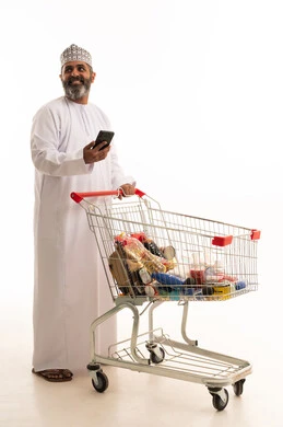 النظر الى شيء ما، تصفح مواقع التواصل الاجتماعي، الاستفادة من الخصومات والتنزيلات، الاستمتاع بالتسوق في المولات والمراكز التجارية، بورتريه لرجل عربي خليجي عماني يرتدي الدشداشة و الكمة يمسك بيدية الجوال و امامة عربة المشتريات، الجسم بالطول الكامل، خلفية بيضاء