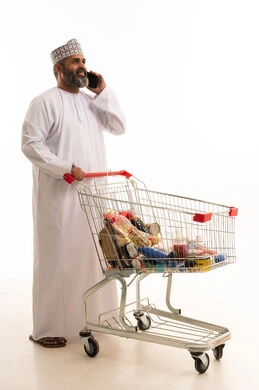 اجراء مكالمة هاتفية عبر الهاتف المحمول، الاستمتاع بالتسوق في المولات والمراكز التجارية، الاستفادة من الخصومات والتنزيلات، بورتريه لرجل عربي خليجي عماني يرتدي الدشداشة و الكمة  يمسك بيدية الجوال و امامة عربة المشتريات، الجسم بالطول الكامل، خلفية بيضاء