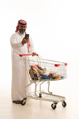 تصفح مواقع التواصل الاجتماعي، الاستفادة من الخصومات والتنزيلات، الاستمتاع بالتسوق في المولات والمراكز التجارية، بورتريه لرجل عربي خليجي سعودي يرتدي الشماغ والثوب التقليدي يمسك بيدية الجوال و امامة عربة المشتريات، الجسم بالطول الكامل، خلفية بيضاء