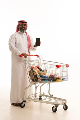 عرض شاشة هاتف محمول سوداء فارغة، الاستمتاع بالتسوق في المولات والمراكز التجارية، الاستفادة من الخصومات والتنزيلات، بورتريه لرجل عربي خليجي سعودي يرتدي الشماغ والثوب التقليدي يمسك بيدية الجوال و امامة عربة المشتريات، الجسم بالطول الكامل، خلفية بيضاء