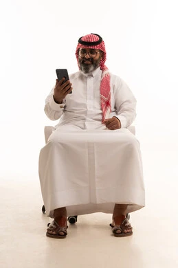 التواصل مع الأهل والاصدقاء، إجراء مكالمة فيديو عبر الجوال، تصفح مواقع التواصل الاجتماعي، استخدام جهاز تقني حديث، بورتريه لرجل عربي خليجي سعودي يرتدي الشماغ والثوب التقليدي يجلس على كرسي يعمل على الهاتف المحمول، خلفية بيضاء