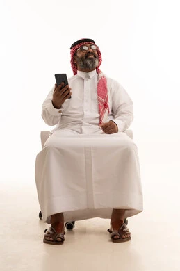 النظر الى شيء ما، التواصل مع الأهل والاصدقاء، إجراء مكالمة فيديو عبر الجوال، تصفح مواقع التواصل الاجتماعي، استخدام جهاز تقني حديث، بورتريه لرجل عربي خليجي سعودي يرتدي الشماغ والثوب التقليدي يجلس على كرسي يعمل على الهاتف المحمول، خلفية بيضاء