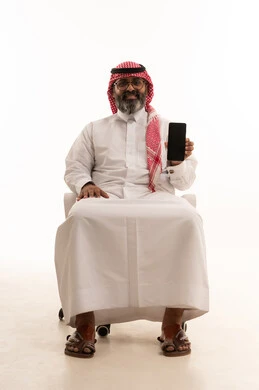 A blank black screen displayed on a mobile phone, browsing social media, using a modern technical device, a portrait of a Saudi Arabian Gulf man wearing a traditional headscarf and thobe sitting on a chair working on a mobile phone, white background.