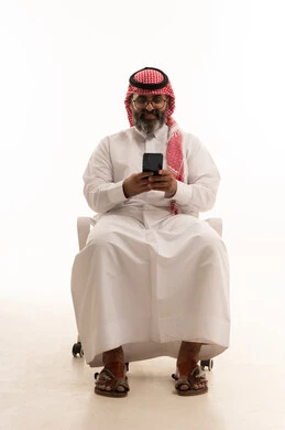 Making a video call via mobile, communicating with family and friends, browsing social media, using modern technology, a portrait of a Saudi Arabian Gulf man wearing a ghutrah and traditional thobe sitting on a chair working on a mobile phone, white background.