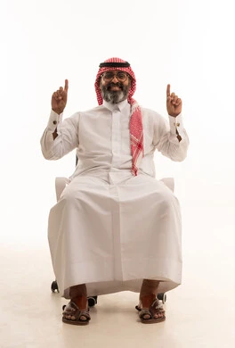 Hand movements and gestures indicating this place (here), pinpointing a specific object, a portrait of a Saudi Arabian Gulf man wearing a ghutrah and traditional thobe sitting on a chair pointing his index finger upwards, with a white background.