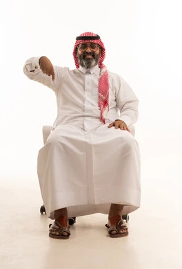 تأكيد حدوث شيء هذه اللحظة, حركات تدل على هذا المكان (هنا)، تحديد موقع معين، بورتريه لرجل عربي خليجي سعودي يرتدي الشماغ والثوب التقليدي يجلس على كرسي يشير بإصابع السبابة نحو الأسفل، خلفية بيضاء