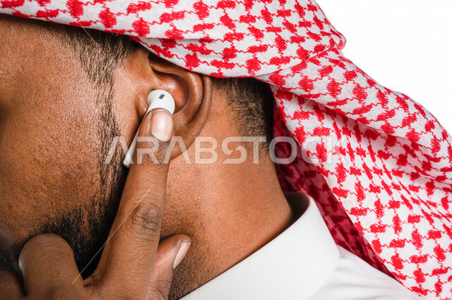 بورتريه بصورة مقربة لرجل عربي خليجي سعودي ، يضع سماعة لاسليكة ، خلفية بيضاء ، سماعة أذن لاسليكة ، مفهوم إستخدام إكسسوارات خاصة بالأجهزة التقنية الحديثة