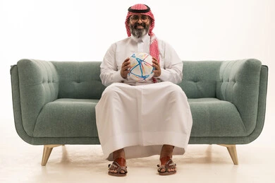 الشغف برياضة كرة القدم ، المباريات والألعاب الرياضية، بورتريه لرجل عربي خليجي سعودي يرتدي الثوب الابيض و الشماغ يجلس على الاريكة يحمل بيده كرة قدم, خلفية بيضاء