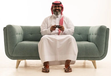 التسلية والترفيه بالفيديو جيمز في وقت الفراغ، استخدام الألعاب الإلكترونية والأجهزة الحديثة، بورتريه لرجل عربي خليجي سعودي يرتدي الثوب الابيض و الشماغ يجلس على الاريكة يمسك عصا التحكم  بتعابير الاندماج و التركيز، خلفية بيضاء