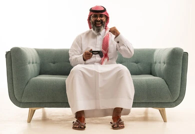 The use of technological devices and modern techniques, spending enjoyable times in recreational video games, a portrait of a Saudi Arabian Gulf man wearing a white thobe and a ghutrah sitting on the sofa holding a game controller, gestures of joy, enjoyment, and immersion, white background.