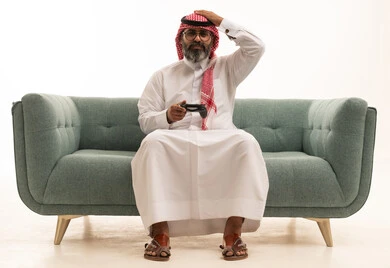Gestures of loss and disappointment, the use of electronic games and modern devices, a portrait of a Saudi Arabian Gulf man wearing a white thobe and a ghutrah sitting on a sofa holding a game controller with his hand on his head showing expressions of regret and sadness, entertainment and leisure with video games in free time, white background.