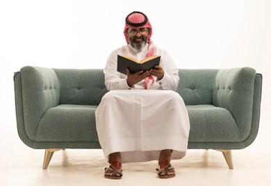 هواية القراءة والاستكشاف، مفهوم التثقيف والبحث العلمي، قضاء وقت الفراغ بالتعلم والثقافة، بورتريه لرجل عربي خليجي سعودي يرتدي الثوب الابيض و الشماغ يجلس على الاريكة يقرأ كتاب، خلفية بيضاء