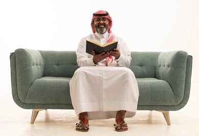 The concept of education and scientific research, spending free time learning and cultural activities, a portrait of a Saudi Arabian Gulf man wearing a white thobe and a shemagh sitting on a sofa reading a book, the hobby of reading and exploration, white background.