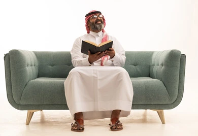 قضاء وقت الفراغ بالتعلم والثقافة، بورتريه لرجل عربي خليجي سعودي يرتدي الثوب الابيض و الشماغ يجلس على الاريكة يقرأ كتاب, مفهوم التثقيف والبحث العلمي، هواية القراءة والاستكشاف، خلفية بيضاء