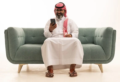 استخدام جهاز تقني حديث ومتطور، العمل عن بعد وانجاز المهام المطلوبة، تصفح مواقع التواصل الاجتماعي، بورتريه لرجل عربي خليجي سعودي يرتدي الثوب الابيض و الشماغ يجلس على الاريكة يمسك بيده الهاتف المحمول، الدردشة والتواصل مع الأهل و الأصدقاء، خلفية بيضاء