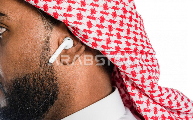 بورتريه بصورة مقربة لرجل عربي خليجي سعودي ، يضع سماعة لاسليكة ، خلفية بيضاء ، سماعة أذن لاسليكة ، مفهوم إستخدام إكسسوارات خاصة بالأجهزة التقنية الحديثة