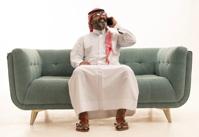 اجراء مكالمة هاتفية عبر الجوال، الدردشة مع الاهل و الاصدقاء، بورتريه لرجل عربي خليجي سعودي يرتدي الثوب الابيض و الشماغ يجلس على الاريكة يحمل الهاتف المحمول، خلفية بيضاء