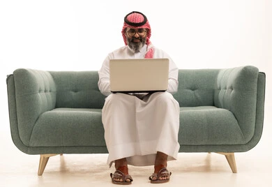 Spending enjoyable times browsing social media, completing work and tasks remotely, a portrait of a Saudi Arabian Gulf man wearing a white thobe and a ghutrah sitting on the couch working on a laptop, conducting online job interviews, white background.