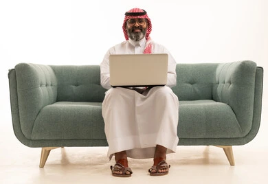 انجاز الأعمال والمهام عن بعد, قضاء أوقات ممتعه بتصفح وسائل التواصل الاجتماعي,بورتريه لرجل عربي خليجي سعودي يرتدي الثوب الابيض و الشماغ يجلس على الاريكة يعمل على الحاسب المحمول, إجراء مقابلات عمل أونلاين, خلفيه بيضاء