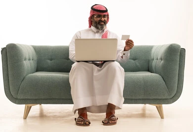 The use of electronic bank cards, the concept of remote payment and settlement through laptop applications, the evolution of technology and technical devices, a portrait of an Arab Gulf Saudi man wearing a white thobe and a shemagh sitting on a sofa working on a laptop, white background.