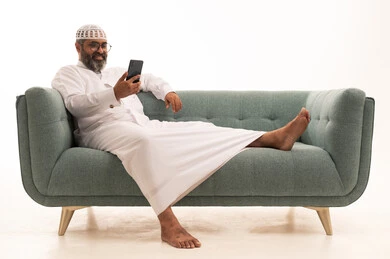 التواصل مع الأهل والاصدقاء، إجراء مكالمة فيديو عبر الجوال، تصفح مواقع التواصل الاجتماعي، استخدام جهاز تقني حديث، بورتريه لرجل عربي خليجي سعودي يرتدي الثوب الابيض و الطاقية يجلس على الاريكة يعمل على الهاتف المحمول، خلفية بيضاء